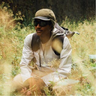 A person crouching in a field, surrounded by tall grass and foliage, dressed in outdoor gear.